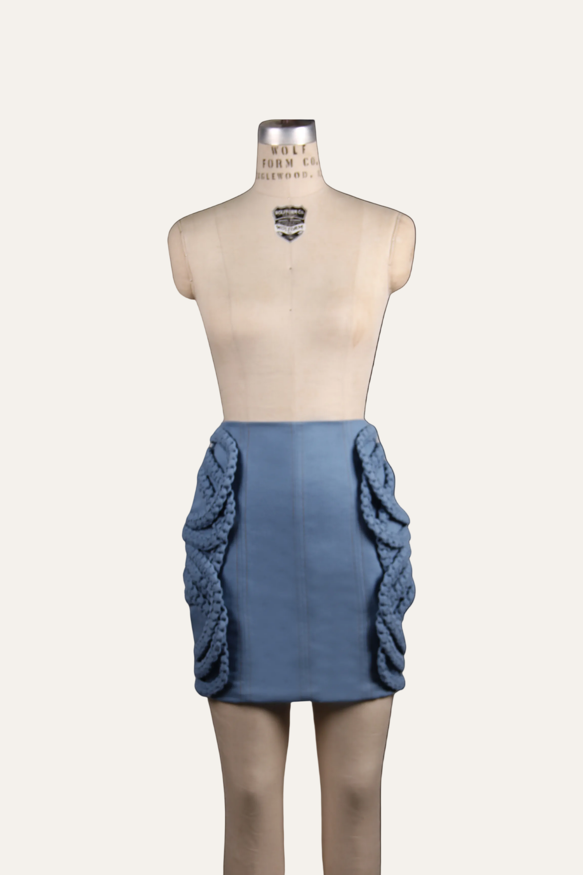 A blue mini skirt with dramatic 3D knotted ruffle details on the sides, displayed on a beige mannequin torso against a light off-white background.